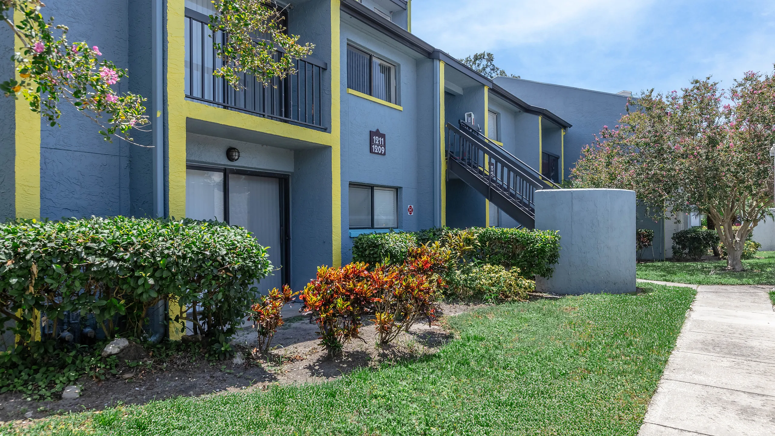 Charming apartment exterior with modern gray and yellow accents, surrounded by lush greenery and vibrant plants.