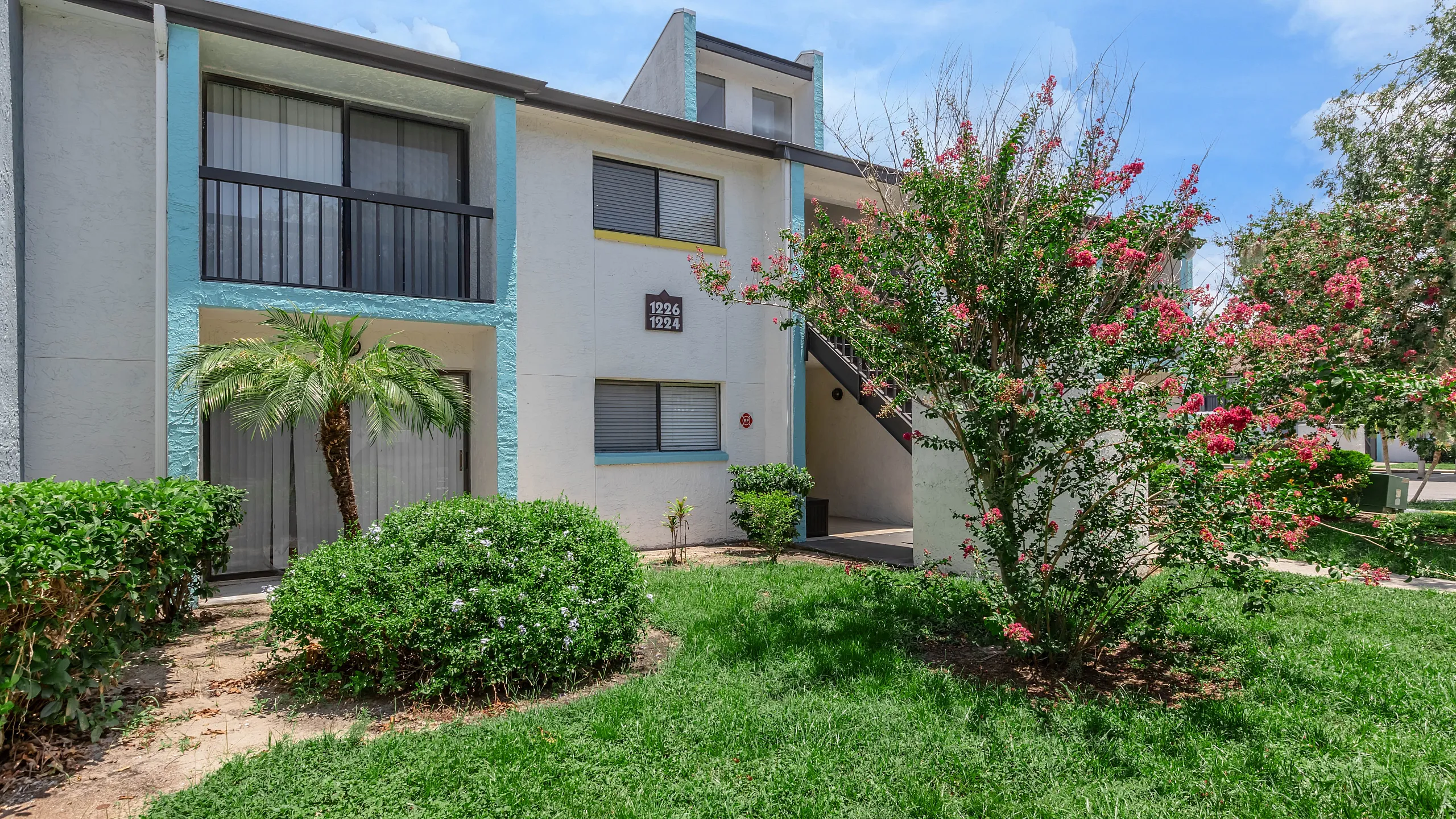 Charming apartment exterior with vibrant landscaping, featuring lush greenery and blooming flowers under a clear blue sky.