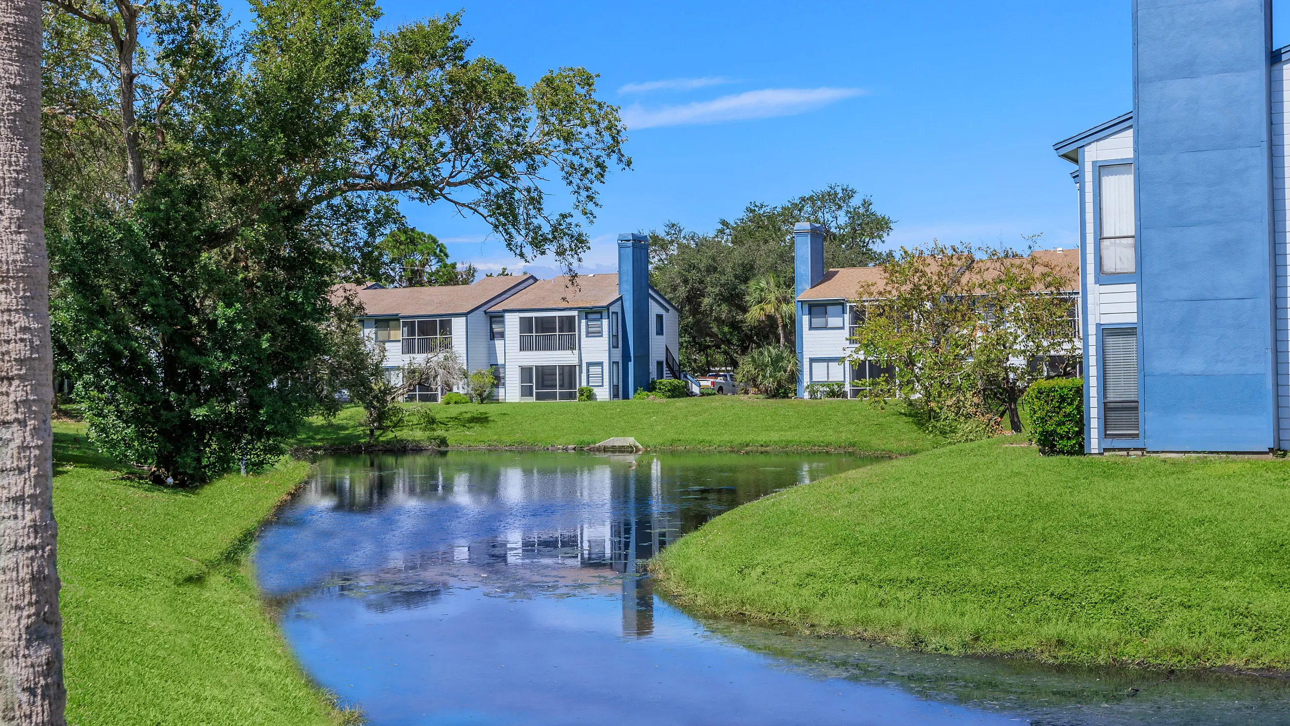 Charming lakeside view of modern blue and white apartment buildings with lush green landscaping, offering serene living in a vibrant community.