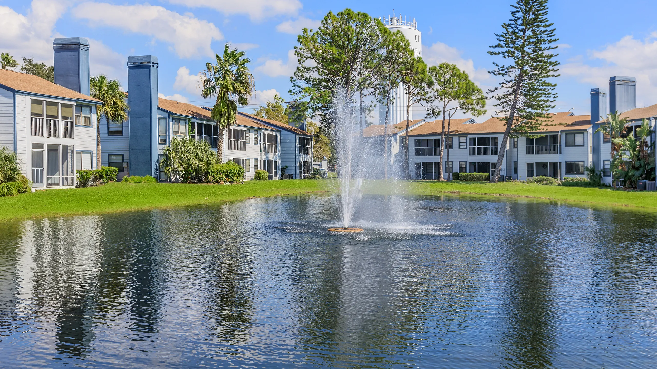 Charming apartment complex with serene landscaped grounds featuring a picturesque pond and fountain, surrounded by lush greenery and modern buildings.