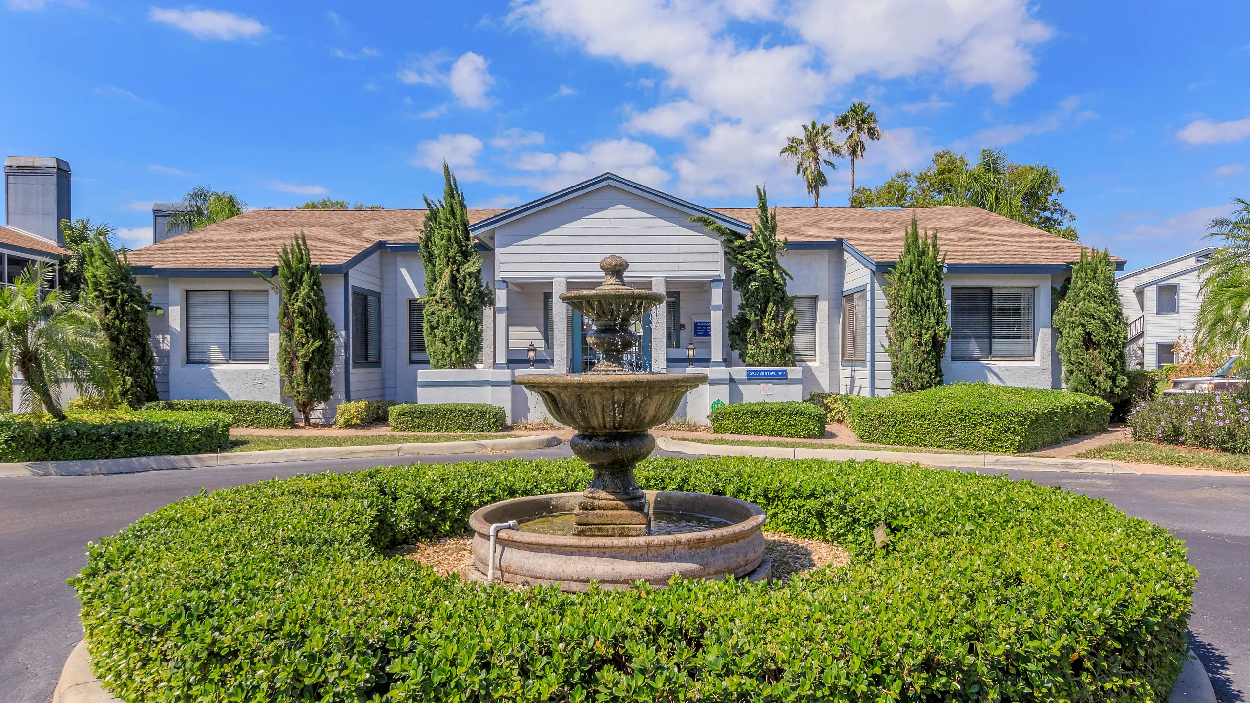 Charming apartment complex exterior featuring a central fountain, lush greenery, and inviting architectural design under a bright blue sky.
