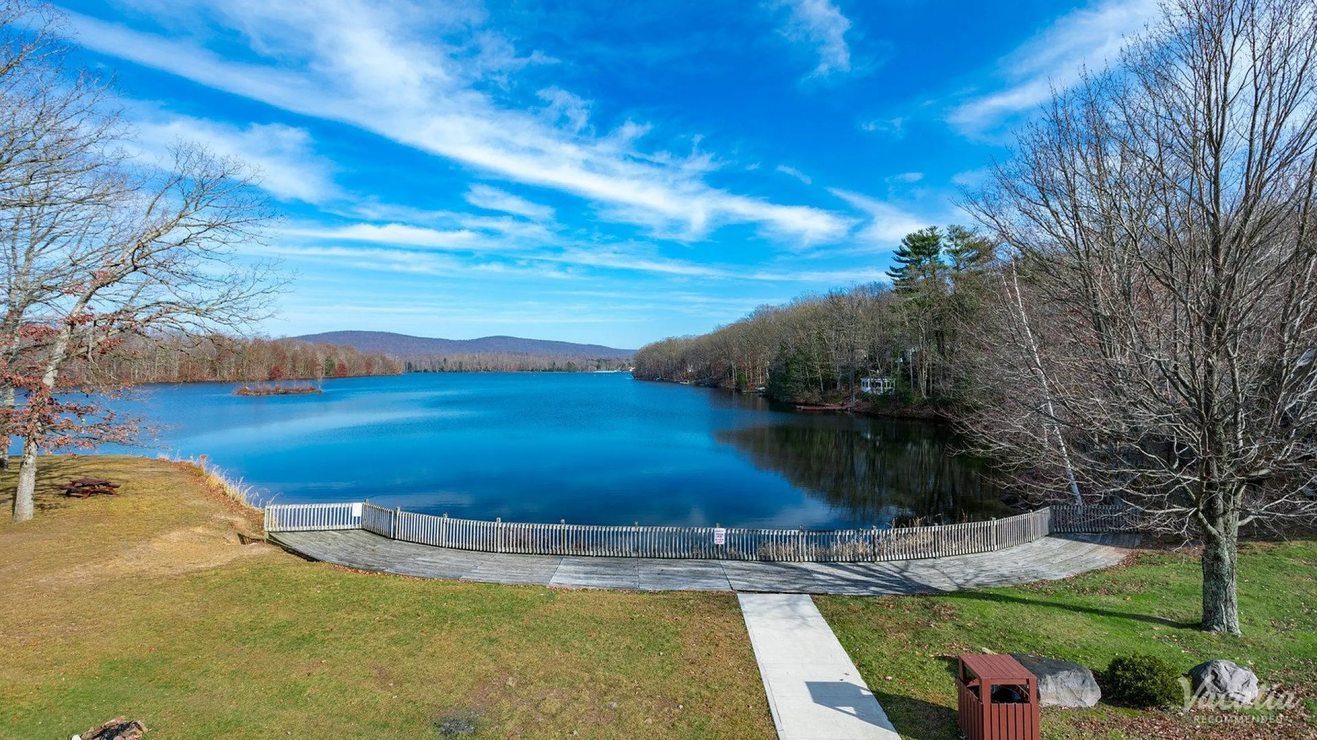 Breathtaking lakeside view with crystal clear water, surrounded by lush greenery and a serene walking path under a bright blue sky.