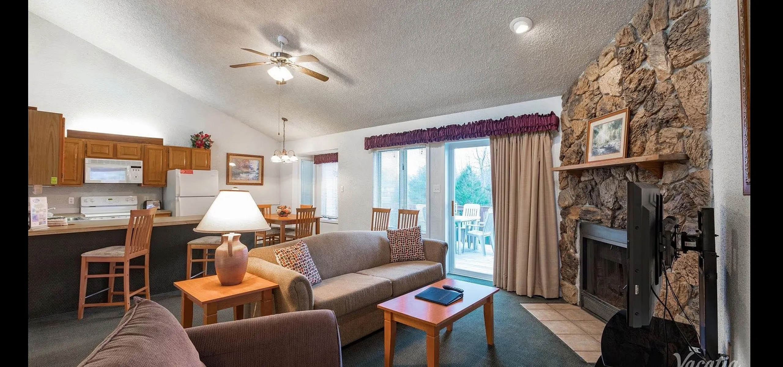 Cozy open-concept living room with stone fireplace, modern kitchen, and sliding glass doors to a scenic patio.