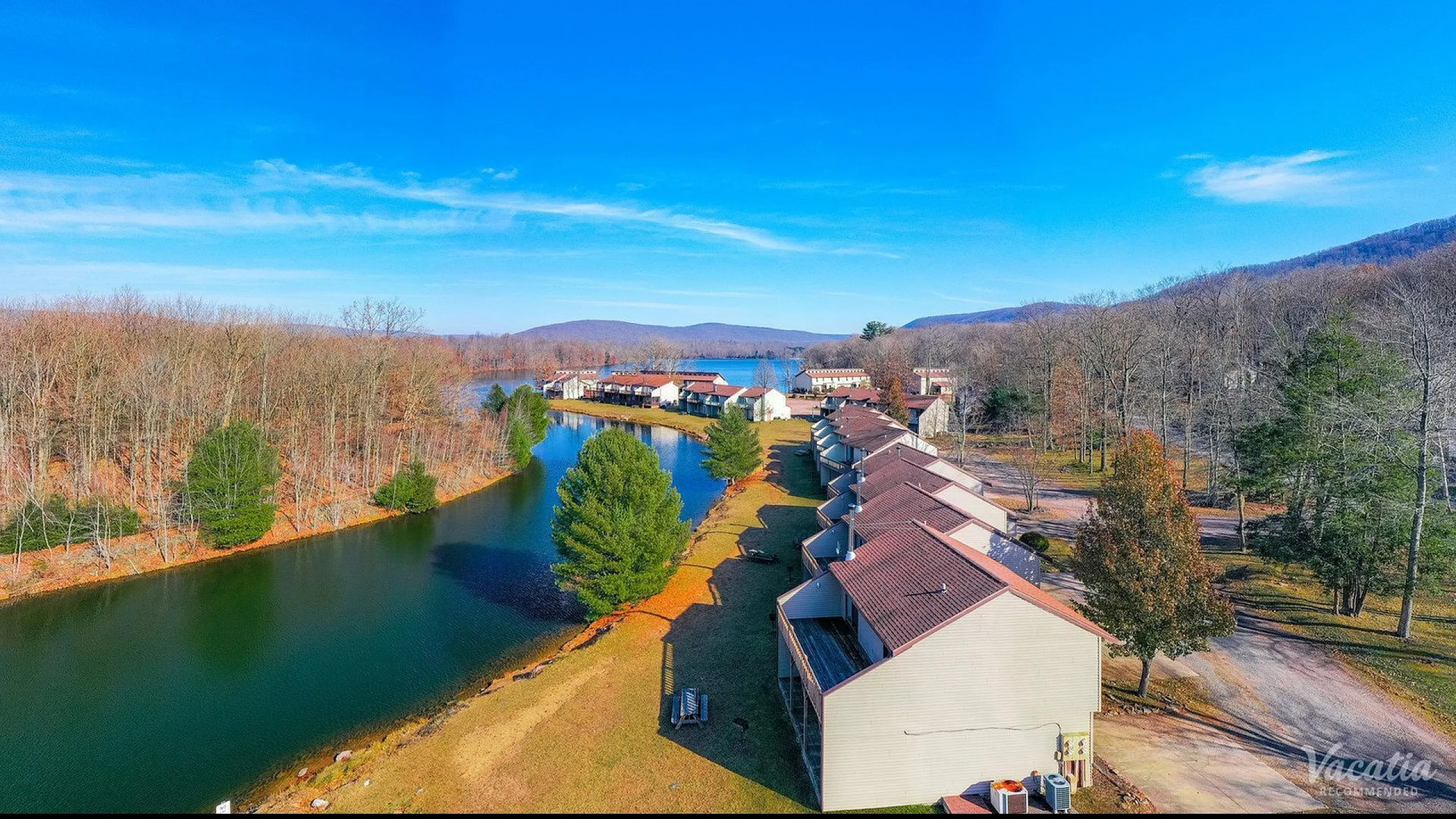 Serene aerial view of a lakeside townhouse community with picturesque wooded surroundings and vibrant blue skies.