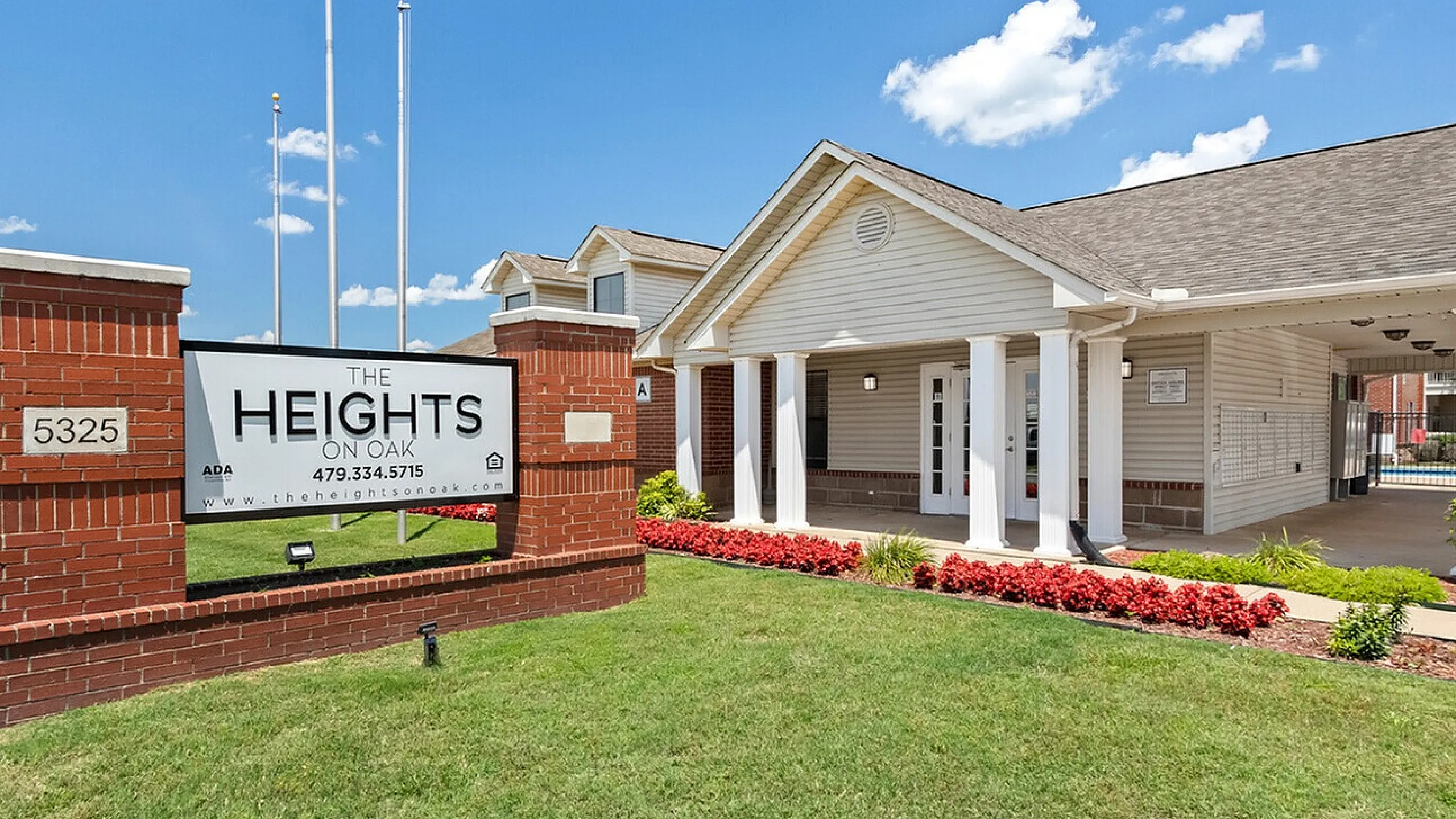Charming entrance of The Heights on Oak with red brick signage, elegant white pillars, and manicured lawns under a clear blue sky.