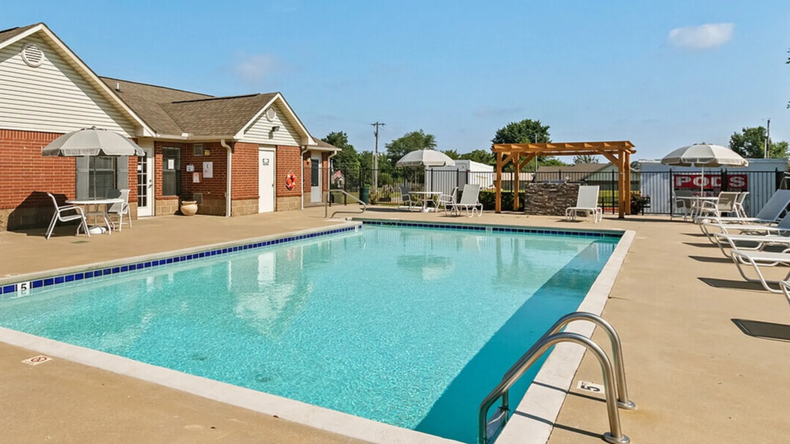 Inviting apartment complex swimming pool area with clear blue water, surrounded by lounge chairs, umbrellas, and a charming brick clubhouse under a sunny sky.