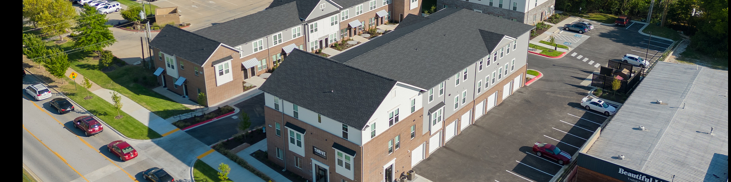 Aerial view of modern townhouse-style apartments surrounded by lush greenery, featuring ample parking and nearby commercial amenities.