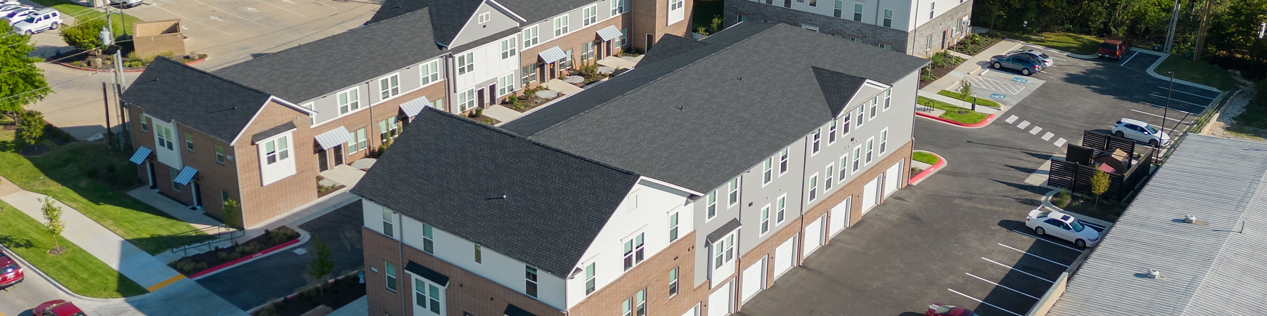 Charming aerial view of a modern apartment complex surrounded by lush greenery and a lively commercial area, offering convenient urban living.