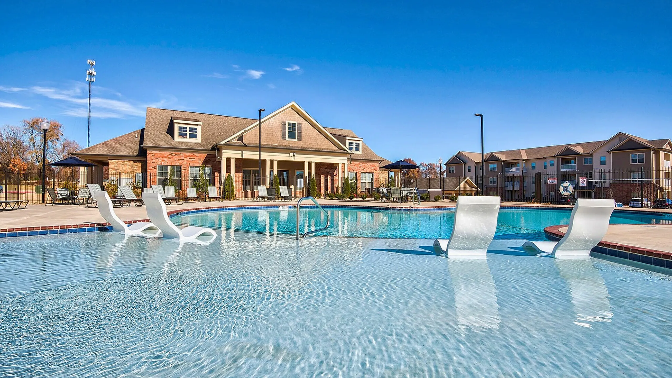 Luxury apartment complex with inviting resort-style pool, sun loungers, and elegant clubhouse under a clear blue sky.