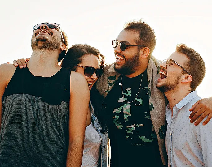 Group of friends laughing together outdoors against a bright, sunny backdrop.