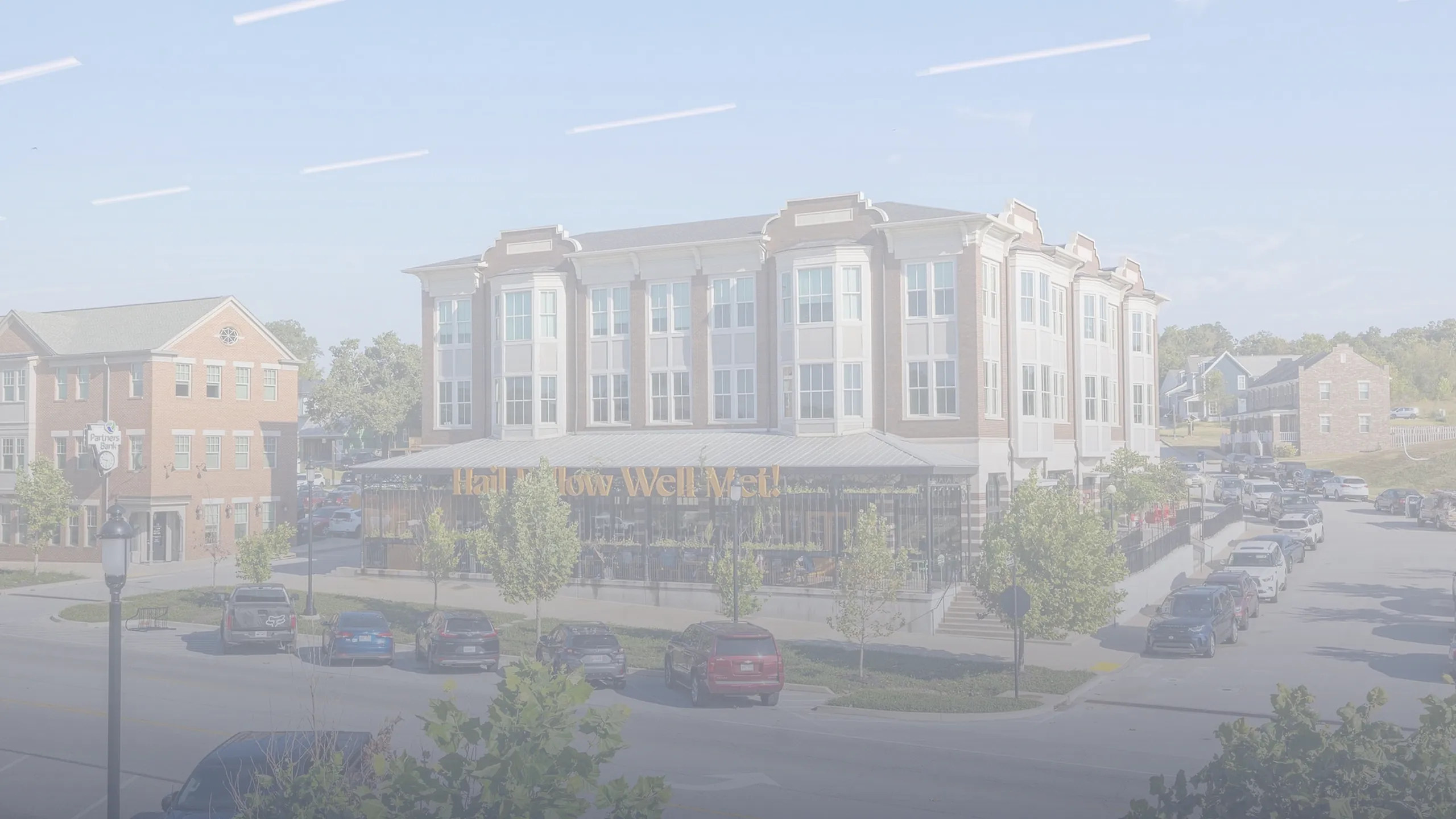 Charming urban street view featuring a stylish three-story building with elegant architecture and inviting storefronts, surrounded by lush greenery and parked cars.
