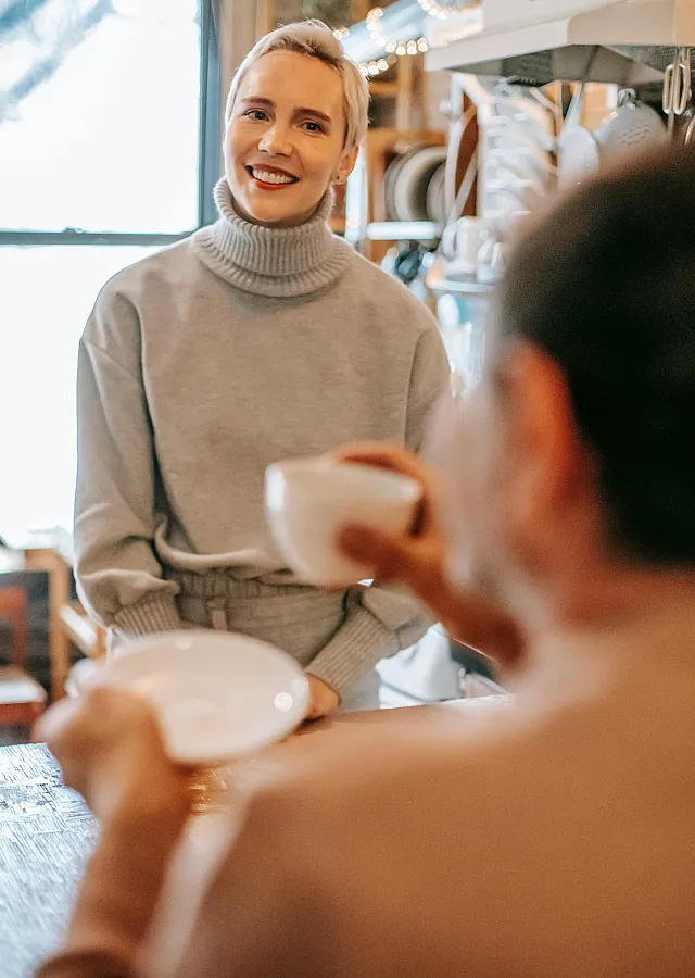 Cozy kitchen scene with a smiling person in a turtleneck sweater, enjoying a warm drink and engaging conversation.