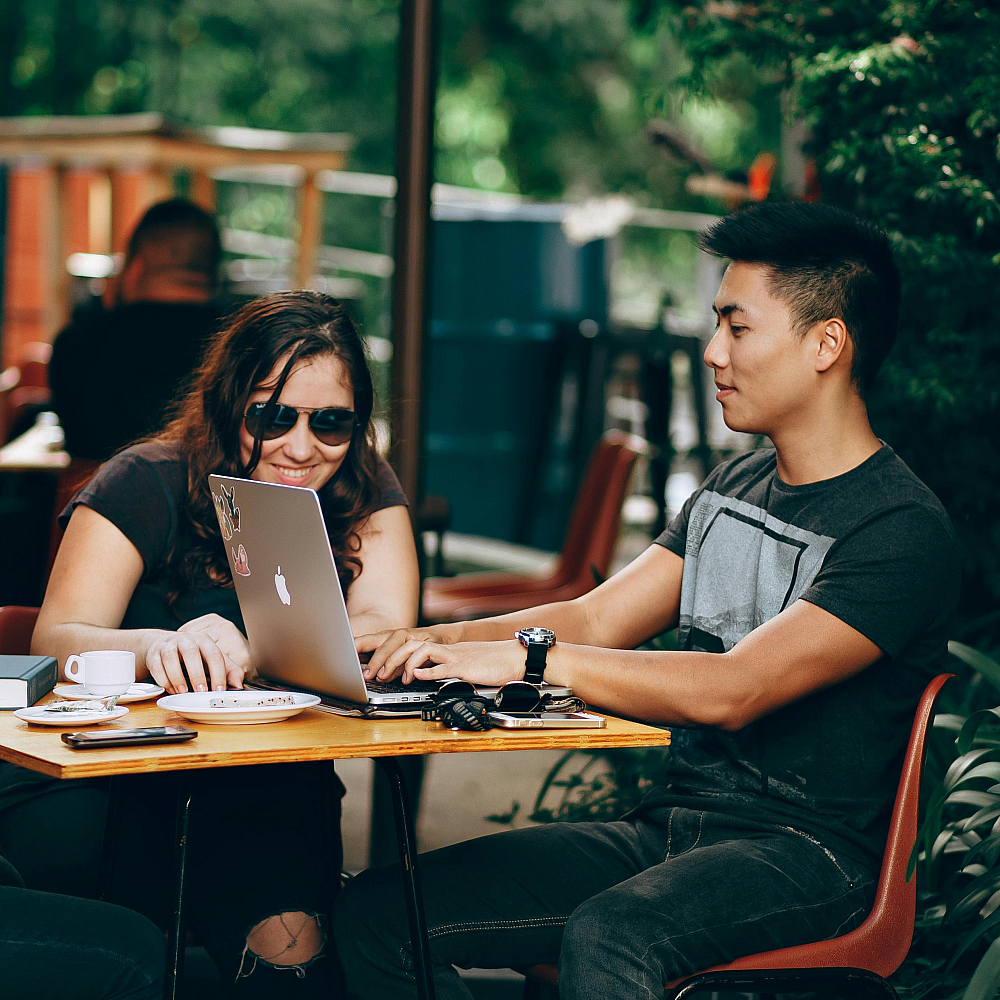 Vibrant outdoor caf&eacute; scene with three friends enjoying coffee and collaborating over a laptop, surrounded by lush greenery.