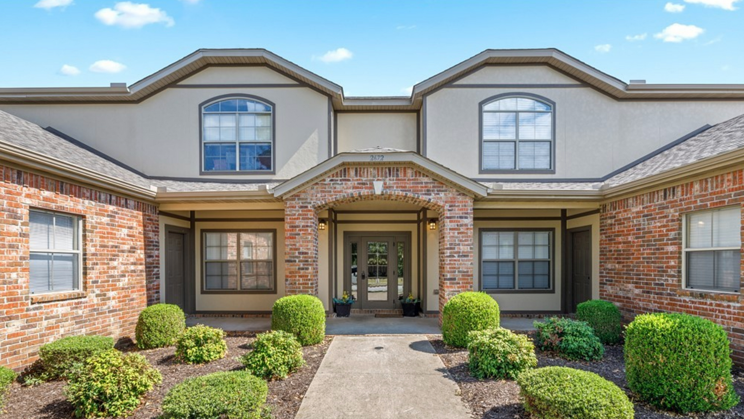 Charming brick apartment exterior with manicured shrubs and inviting entrance under a bright blue sky.