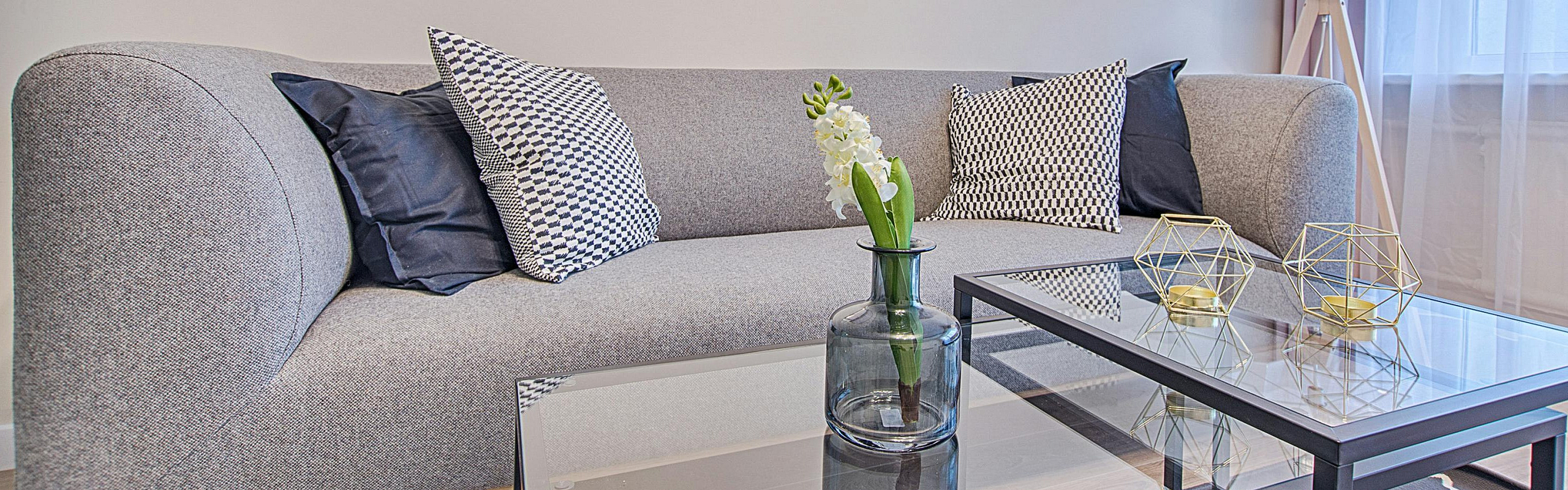 Chic living room with a modern gray sofa, stylish geometric pillows, glass coffee tables, and elegant decor under soft natural light.