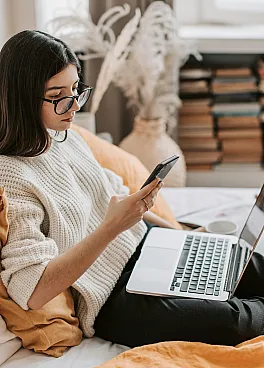 Cozy living space with a person relaxing on a comfortable bed, working on a laptop and phone, surrounded by warm colors and natural light.
