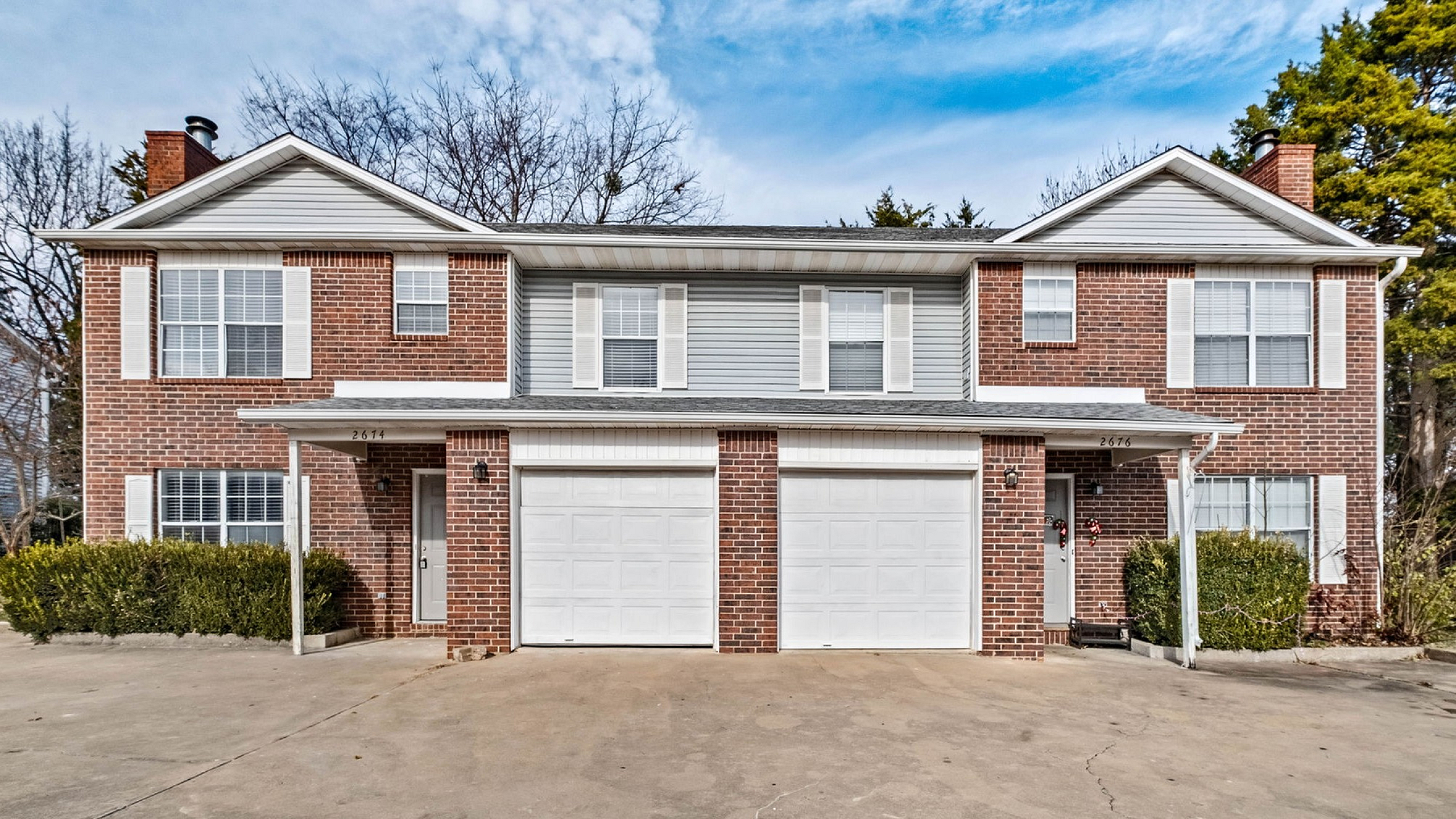 Charming brick duplex with two white garage doors, surrounded by lush greenery and a spacious driveway, set against a vibrant blue sky.
