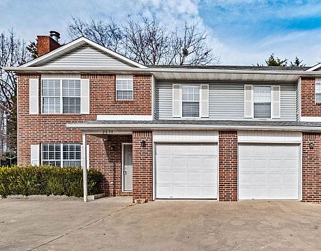 Charming duplex with elegant brick facade, spacious garages, and inviting front porches surrounded by greenery.