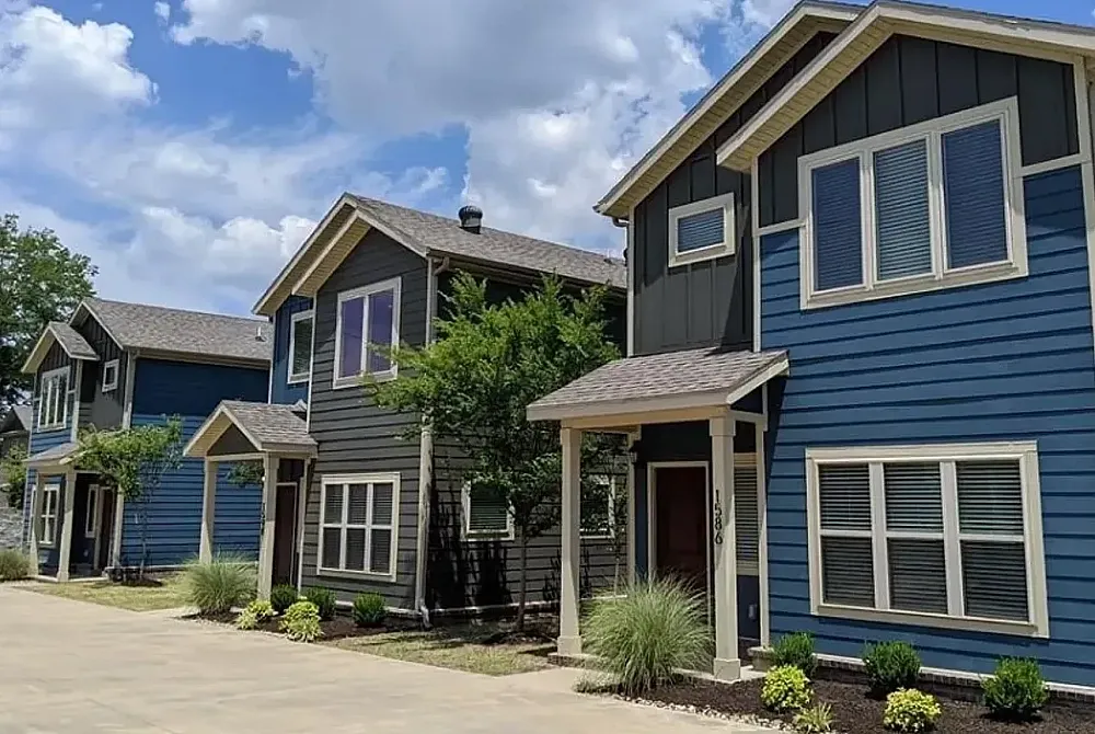 Charming two-story beige apartment complex with rustic stonework, surrounded by mature trees and a classic street lamp.