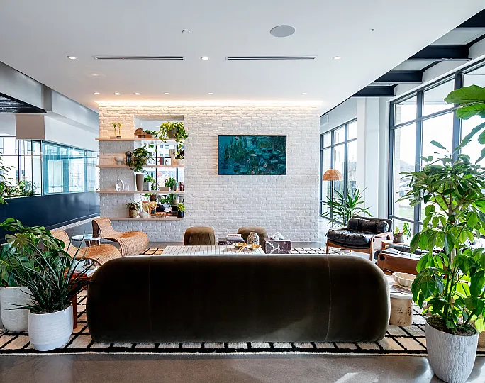 Modern living room with a stylish sofa, lush indoor plants, large windows, and elegant shelving on a white brick wall.