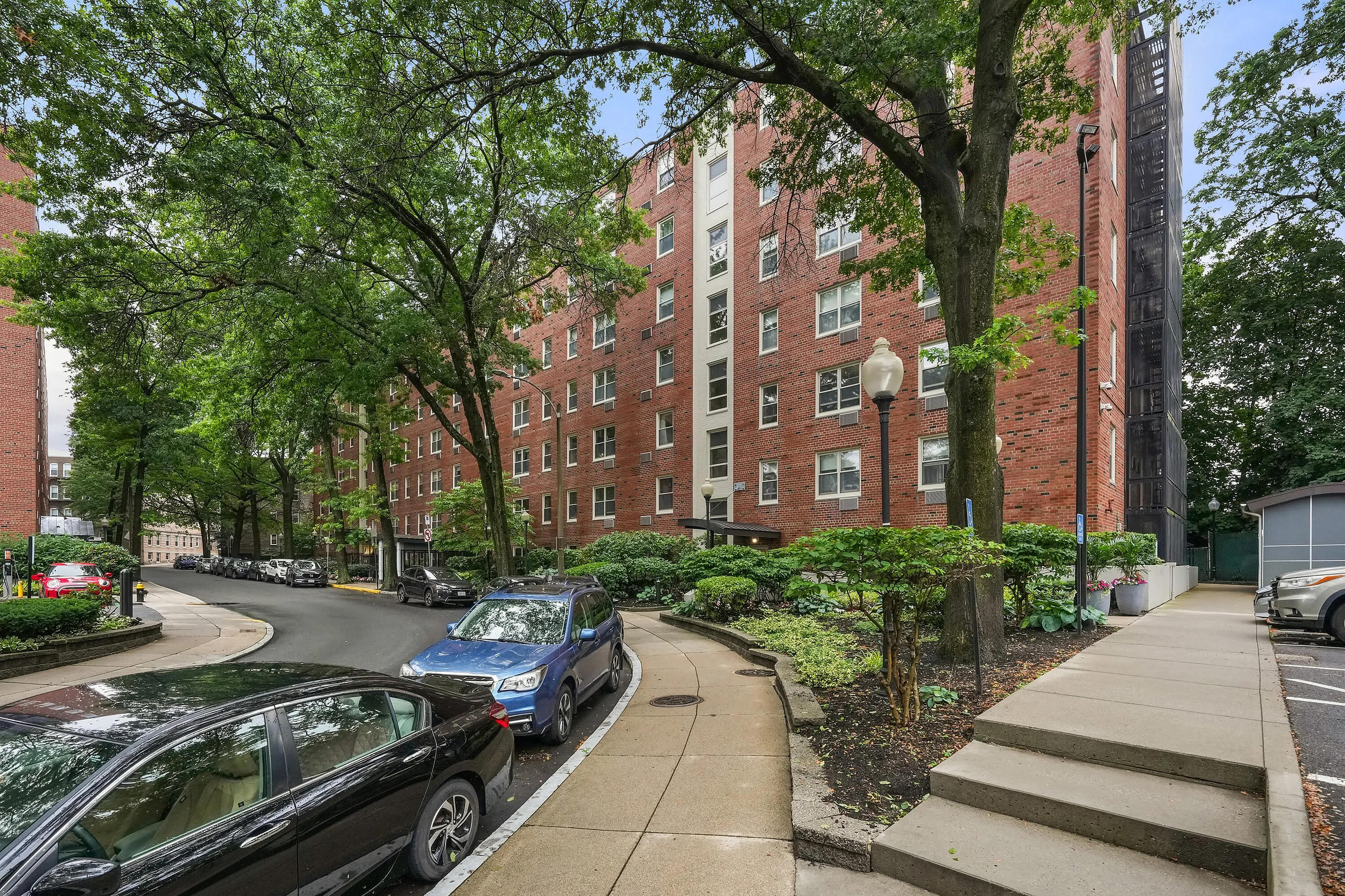 Charming brick apartment building surrounded by lush green trees and a well-maintained pathway, offering a serene urban living experience.