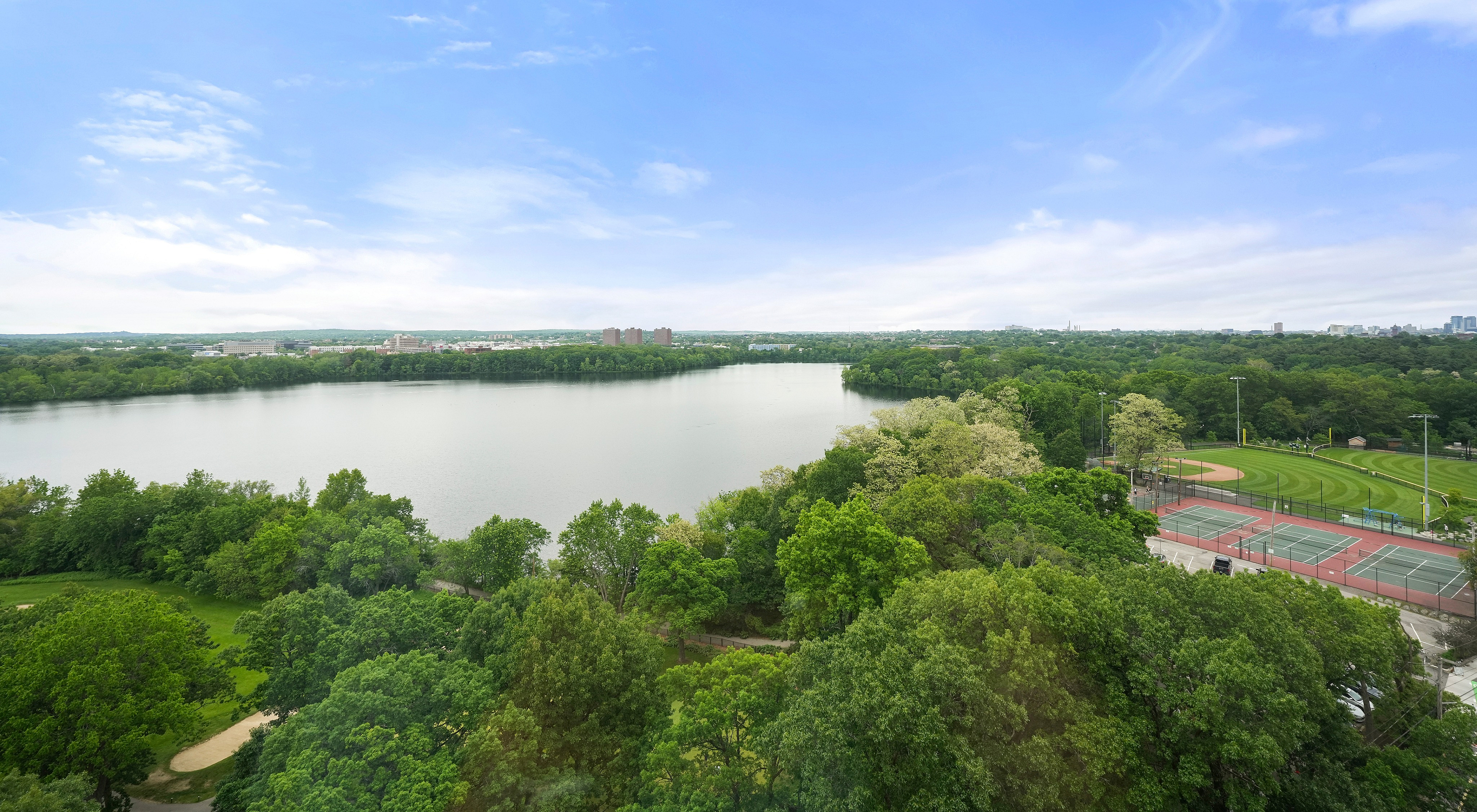Breathtaking aerial view of lush greenery surrounding a serene lake, with sports courts providing recreational charm under a clear blue sky.