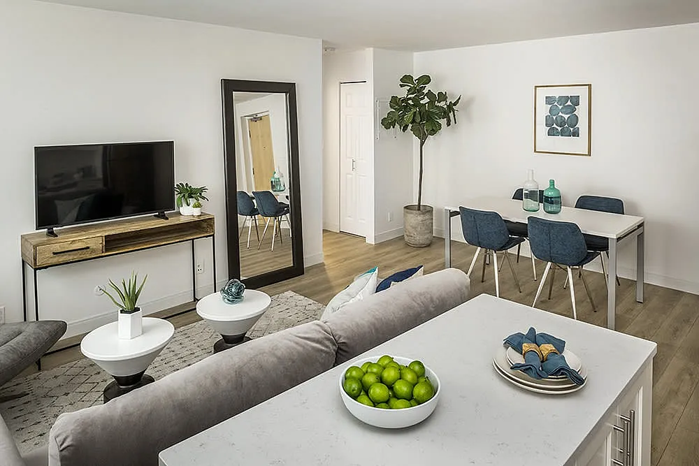 Modern living room with sleek decor, featuring a comfortable gray sofa, stylish coffee tables, and a large mirror enhancing the open space. A chic dining area with blue chairs and elegant art add sophistication.