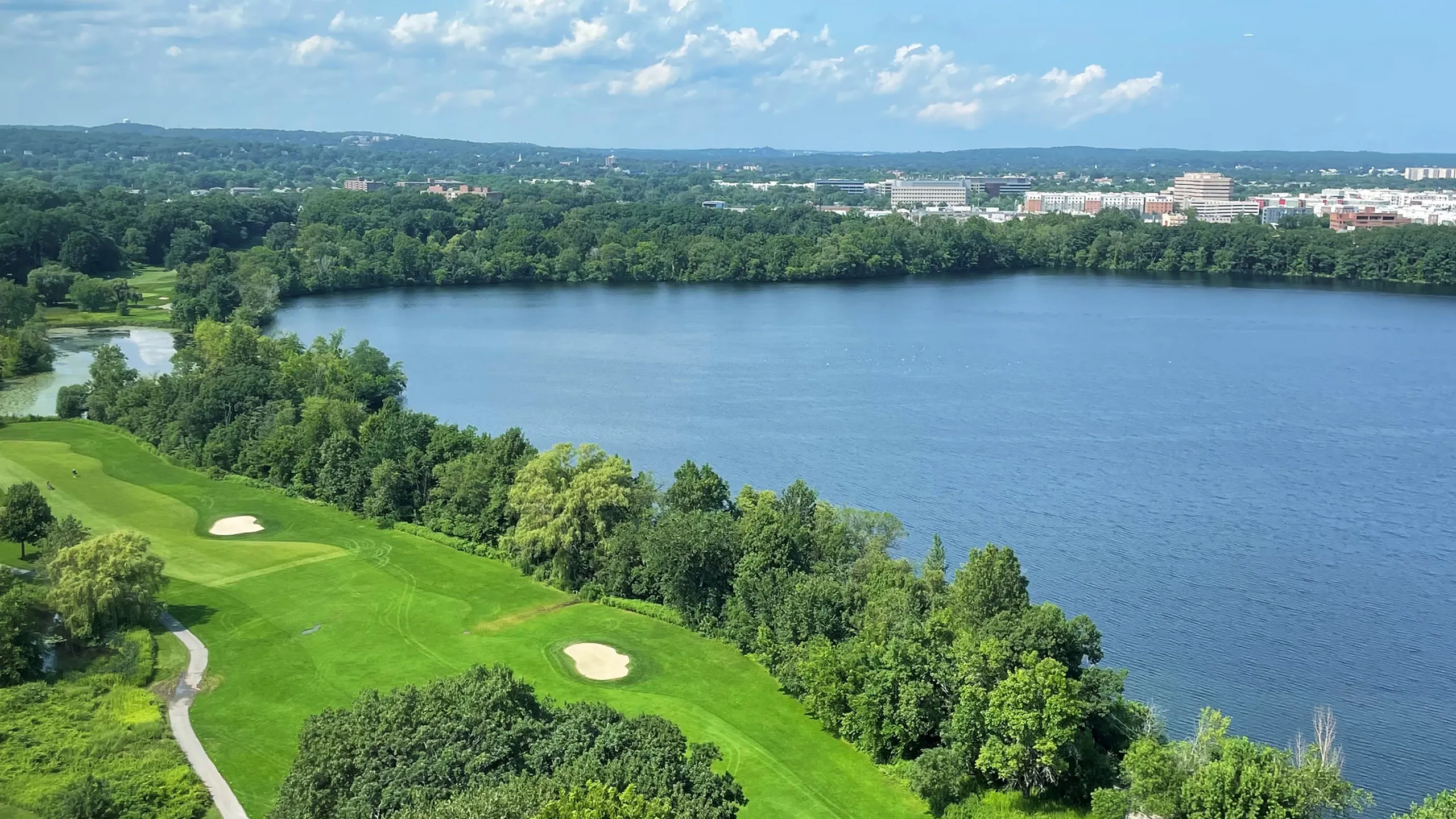 Scenic view of a lush green golf course beside a tranquil lake, surrounded by verdant forests and a distant city skyline under a blue sky.