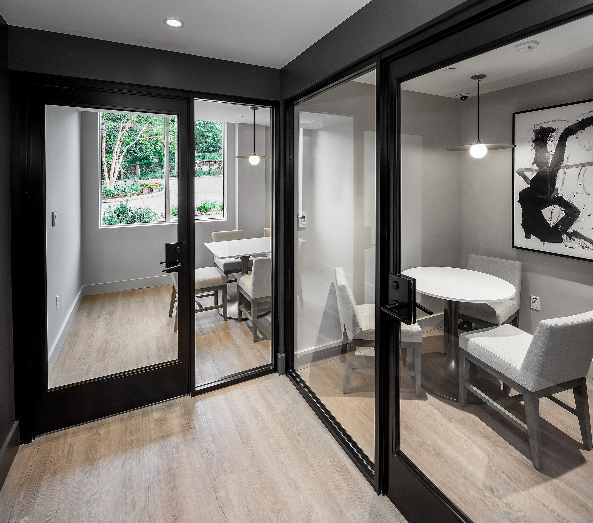 Elegant apartment space featuring two enclosed seating areas with chic glass doors, modern decor, and natural light streaming through a large window with a view of lush greenery.