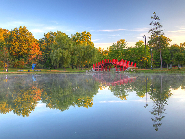 Tranquil park scene with a vibrant red bridge reflected in a serene pond surrounded by lush green trees.