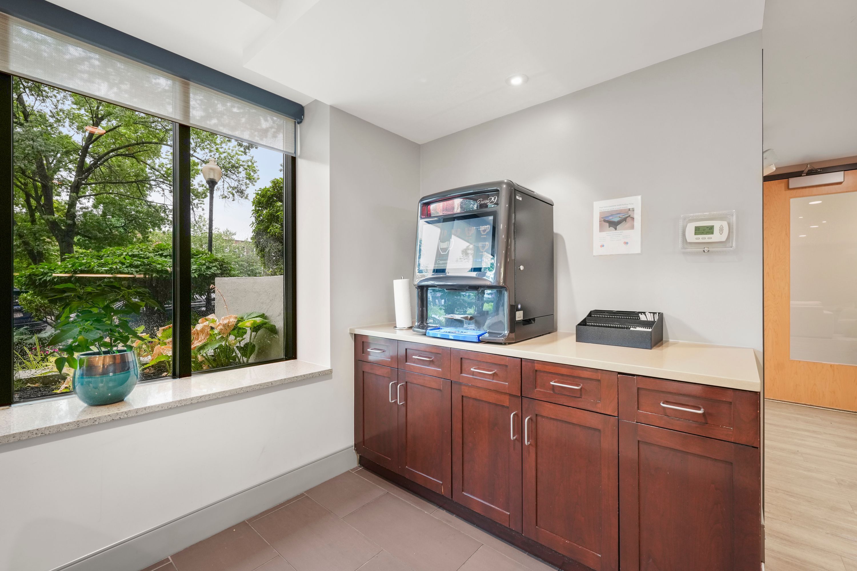 Bright community kitchen corner with large windows overlooking lush greenery, featuring modern cabinetry and a sleek water dispenser.
