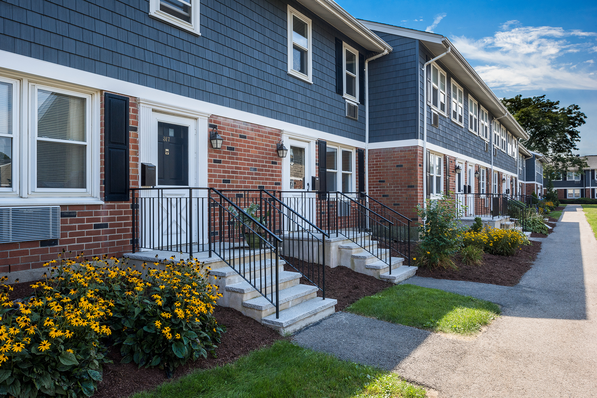 Charming townhouse facade with elegant brickwork, neatly trimmed hedges, and vibrant flowerbeds under a clear blue sky.