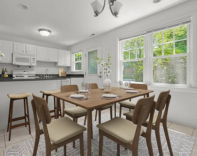 Bright and inviting dining area with a wooden table set for six, large windows overlooking lush greenery, and a modern kitchen with a breakfast bar and stylish lighting.