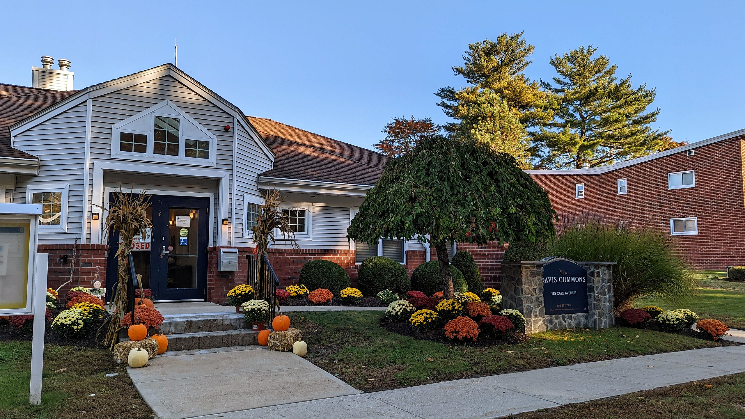 Charming residential building with inviting entrance, seasonal decorations of pumpkins and colorful flowers, complemented by lush greenery under a clear blue sky.