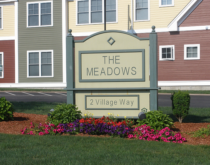 Welcoming sign at The Meadows with vibrant flower bed in front, set against colorful apartment buildings.
