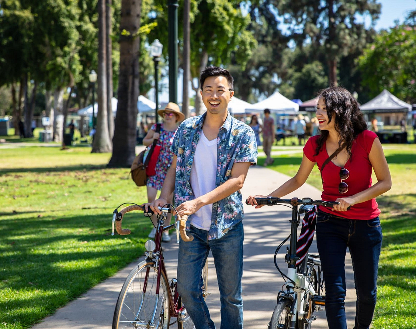 Joyful couple strolling with bicycles in a sunlit park, surrounded by lush green trees and market tents, radiating a vibrant summer atmosphere.