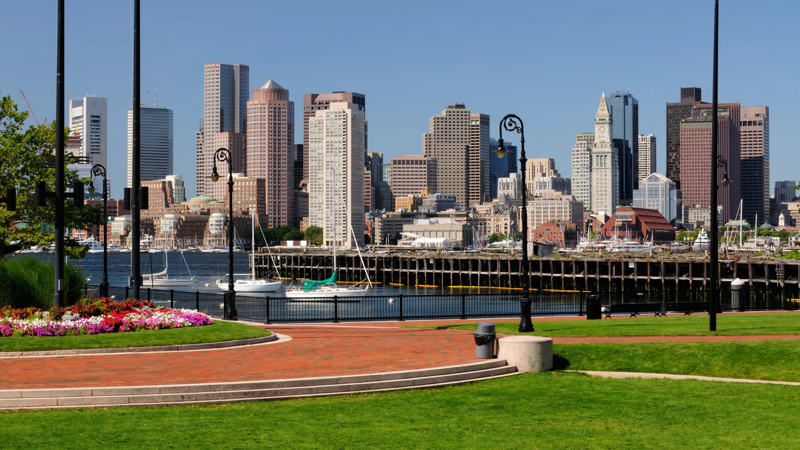 Vibrant cityscape view with lush green park and colorful flowerbeds by the waterfront, set against a backdrop of modern skyscrapers and a clear blue sky.
