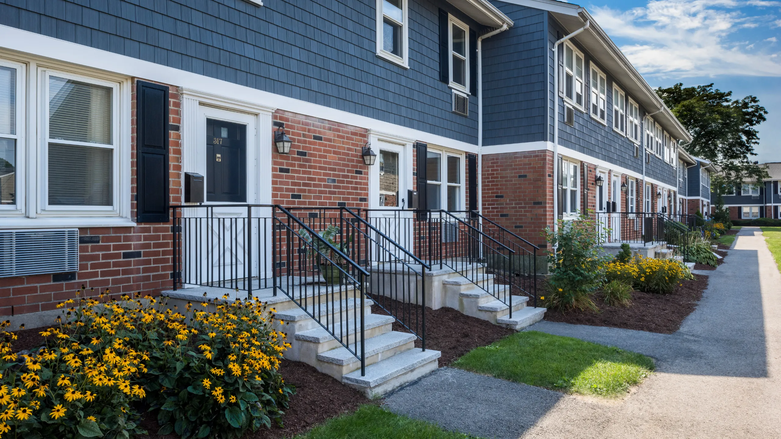 Charming brick townhouse exterior with elegant black shutters and inviting front steps, flanked by vibrant yellow flowers and well-maintained greenery.