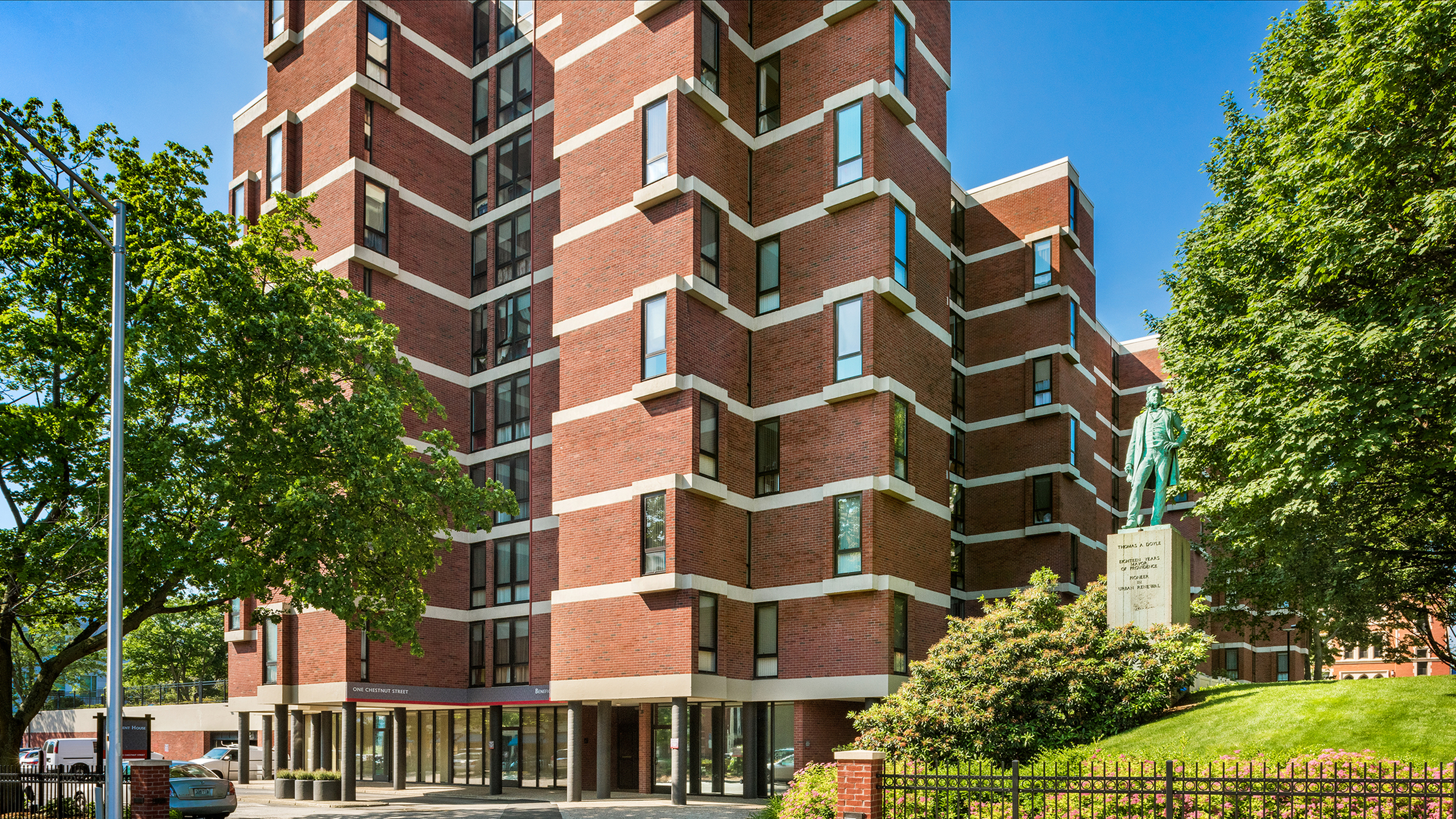 Modern red brick apartment building with large windows, surrounded by lush greenery and a spacious parking area, under a clear blue sky.