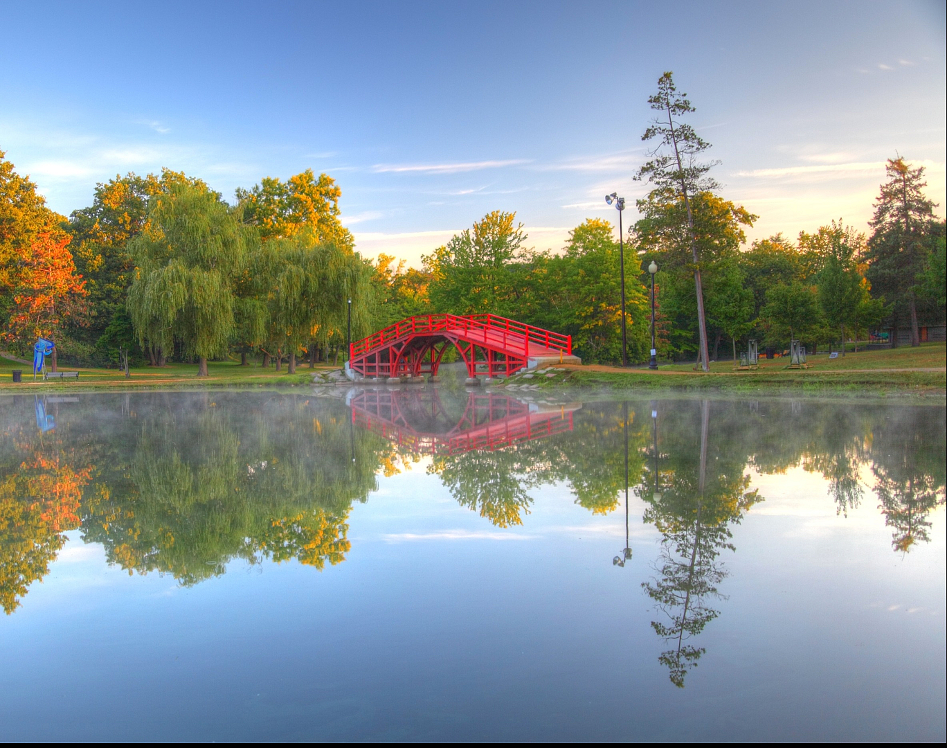 Serene park scene featuring a charming red bridge over a tranquil pond, surrounded by lush, colorful trees reflecting in the water.