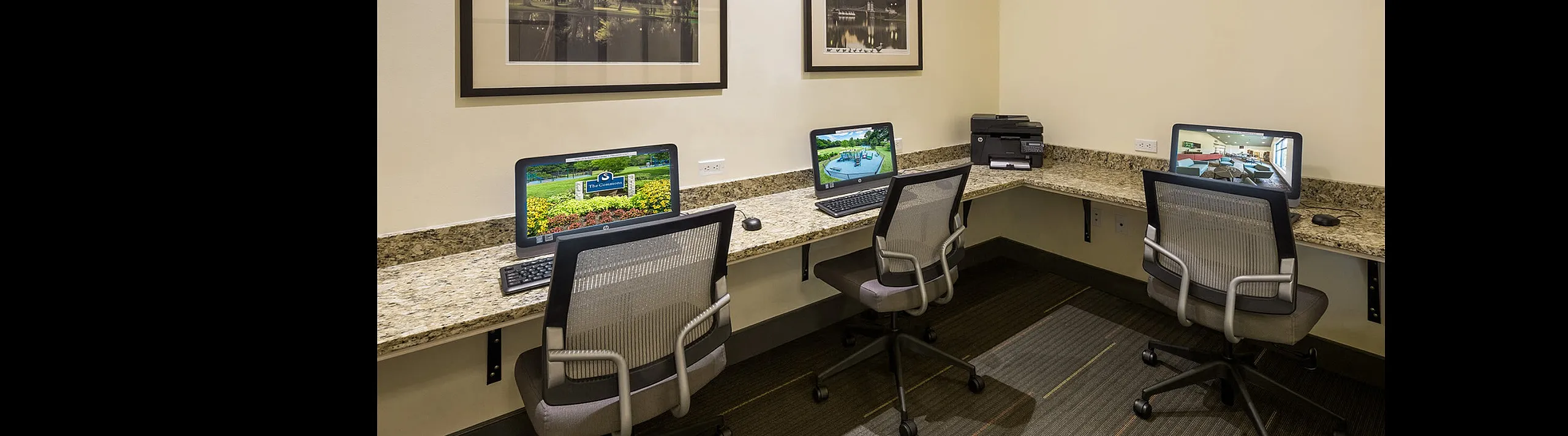 Modern business center featuring three computers on a granite countertop with elegant framed artwork and plush office chairs.