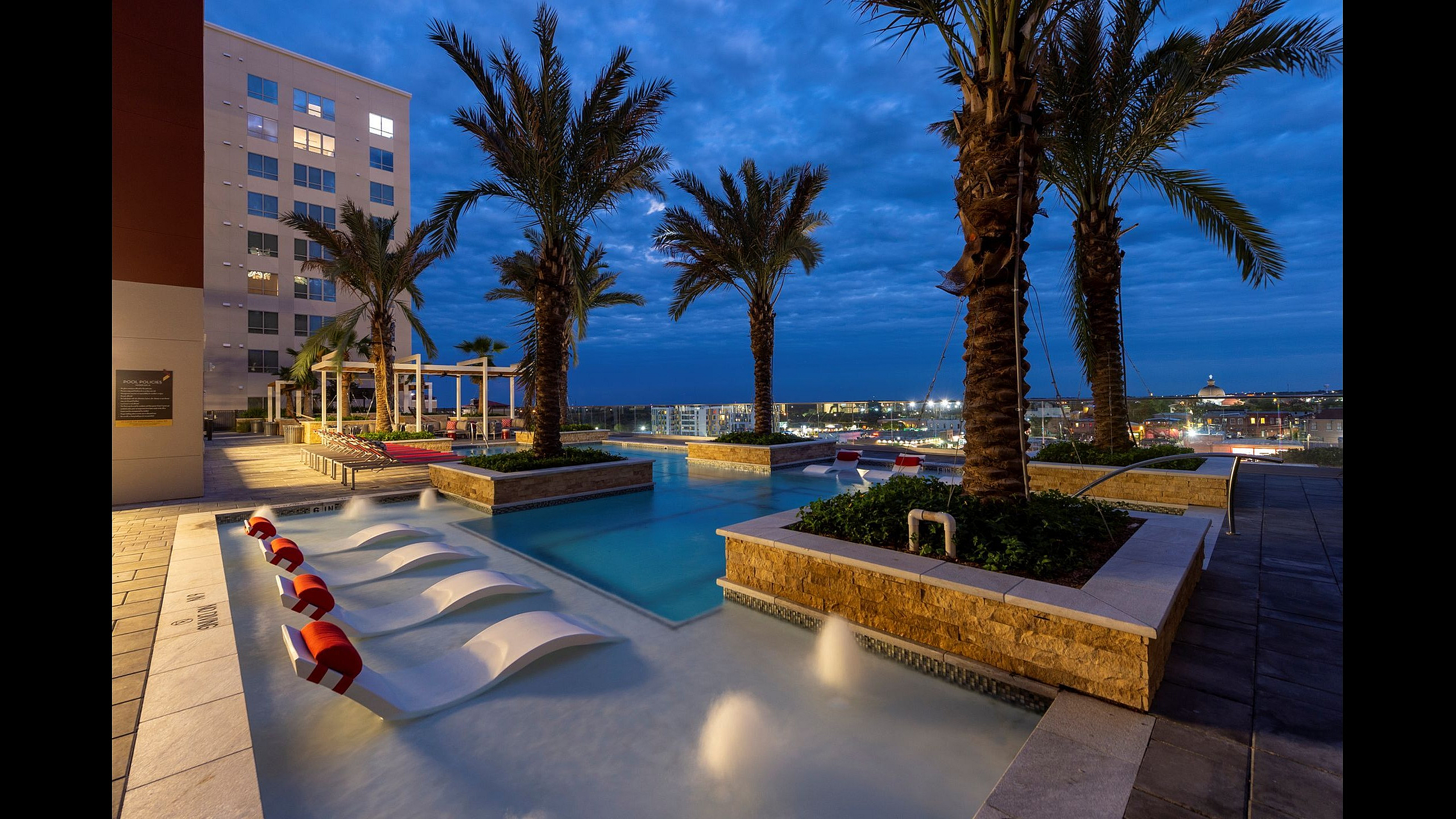 Luxurious rooftop pool area at dusk featuring elegant lounge chairs and palm trees, offering stunning city views.