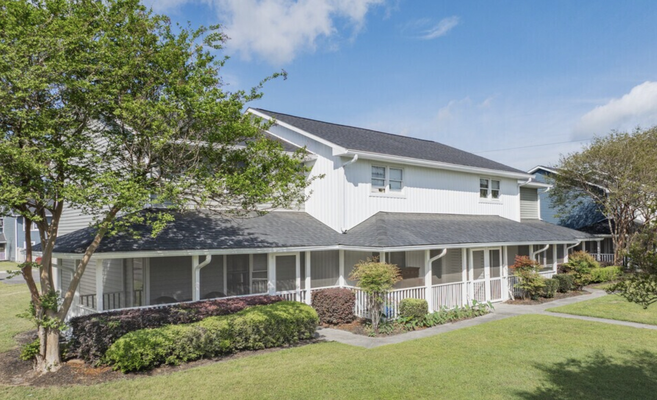 Charming two-story home with a classic white exterior and cozy front porch, nestled among lush green gardens and mature trees, perfect for tranquil living.