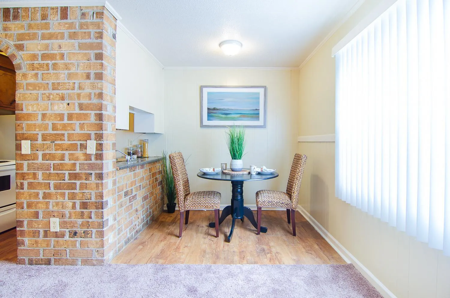 Charming dining area with a round table set for two, featuring wicker chairs and a decorative plant, surrounded by warm brick and a soft beige palette.