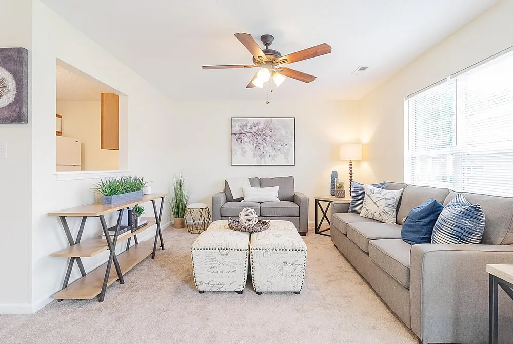 Bright and inviting living room with plush gray sofas, stylish ottomans, decorative cushions, and a ceiling fan, creating a cozy ambiance for relaxation.