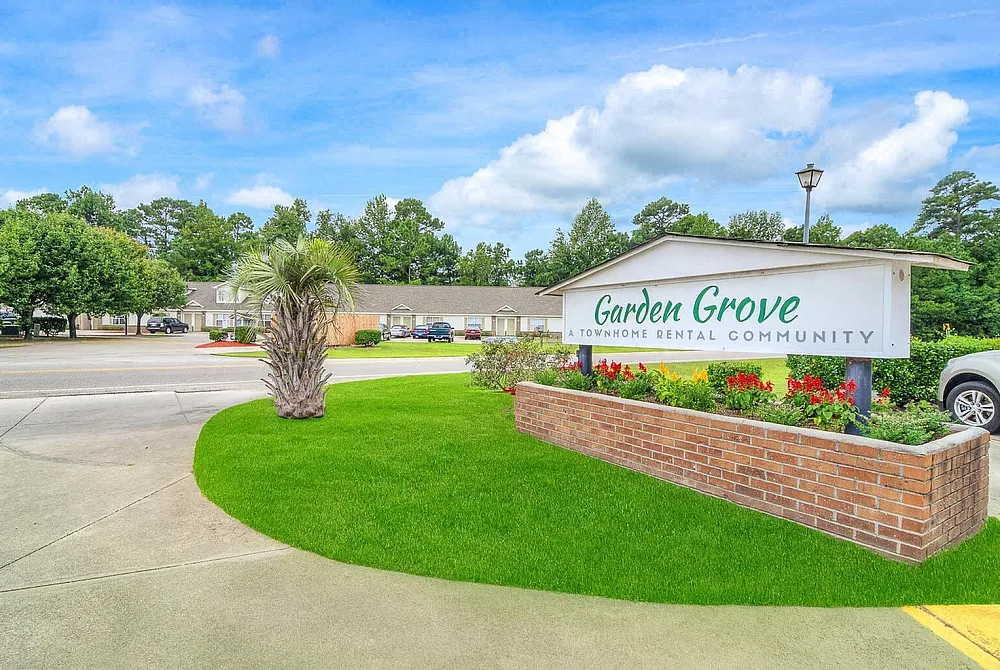 Inviting entrance to Garden Grove Townhome Rental Community surrounded by lush green lawns and vibrant flowers under a clear blue sky.