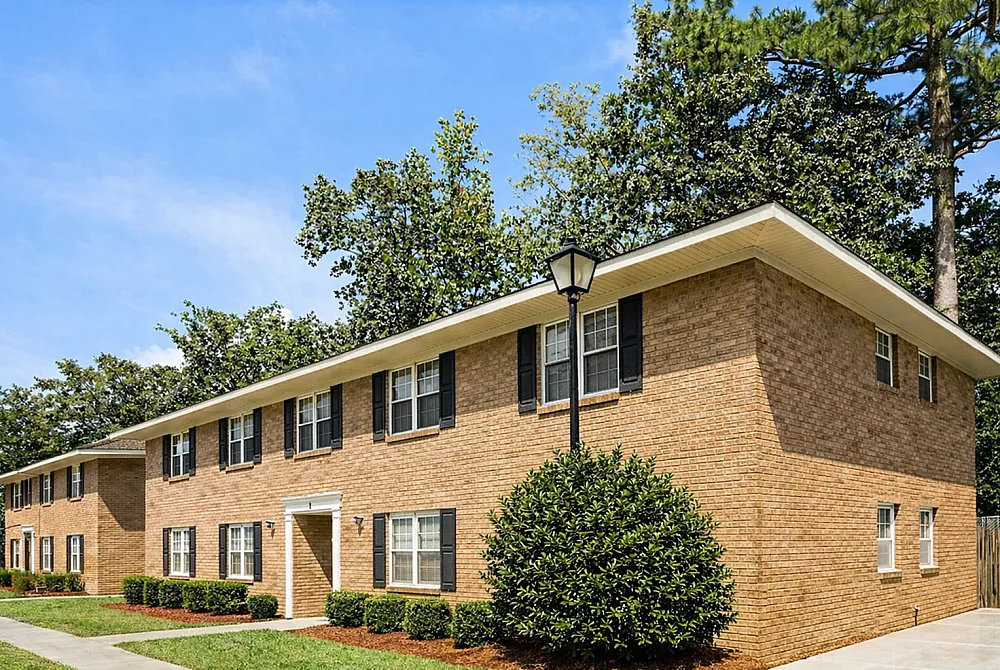 Charming brick apartment building surrounded by lush greenery and blue skies, featuring classic black shutters and a welcoming entrance.