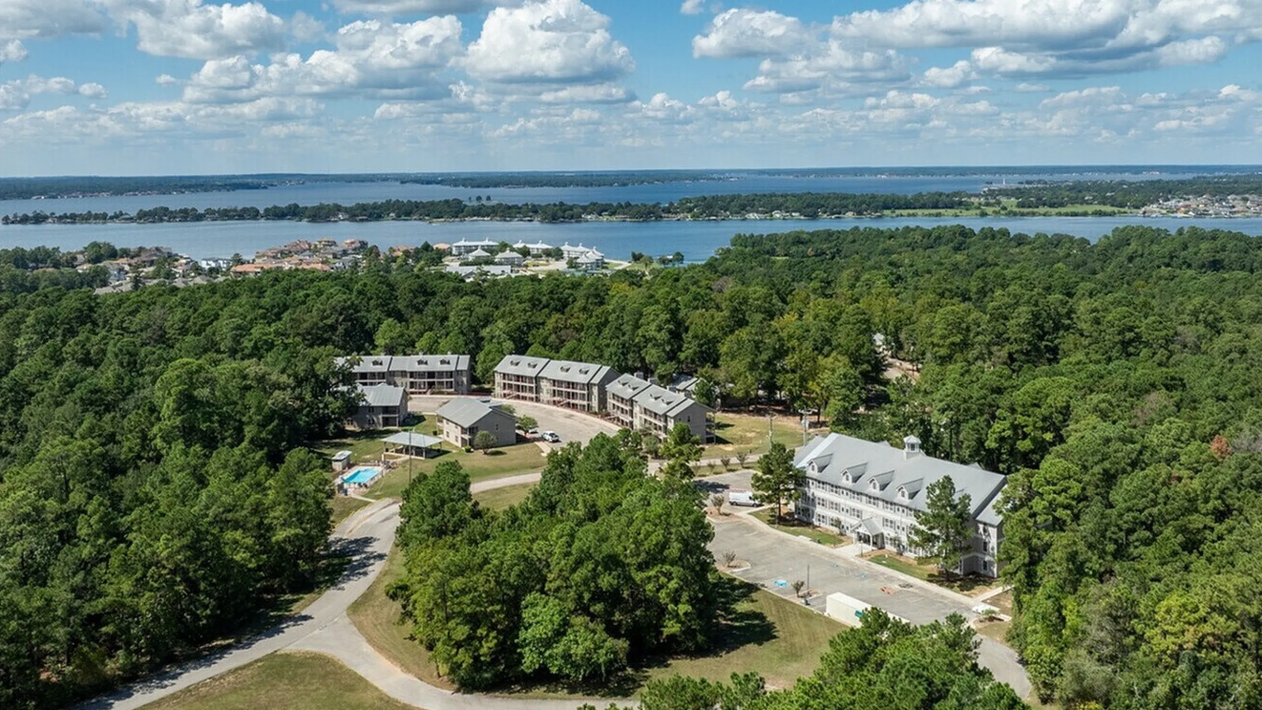 Scenic aerial view of a picturesque waterfront apartment complex surrounded by lush greenery, with a serene lake and expansive landscapes under a vibrant blue sky.