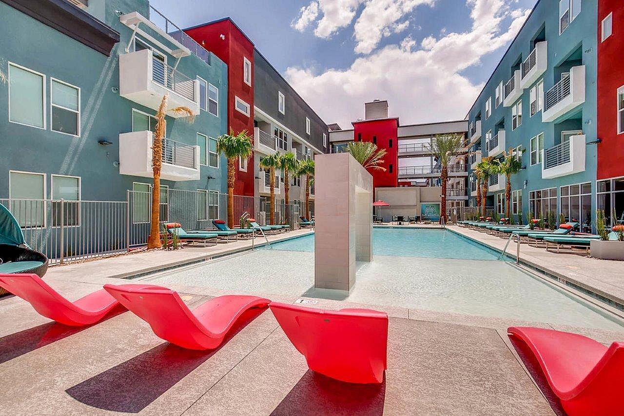 Modern apartment complex with a vibrant exterior showcasing a luxurious swimming pool, stylish red lounge chairs, and palm trees under a bright blue sky.
