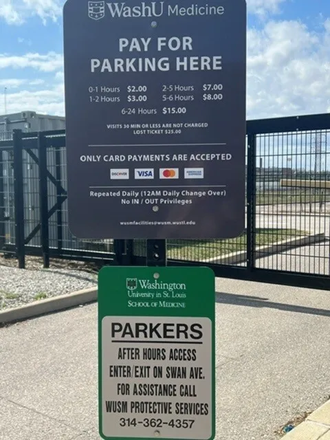 Parking information sign at WashU Medicine with rates and payment options, located in front of a gated facility under a clear blue sky.